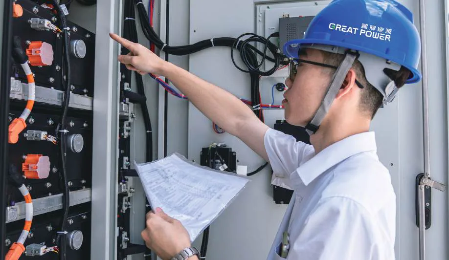 Technician wearing a blue helmet marked “鹏辉能源 GREAT POWER” inspecting an open energy storage cabinet with drawings.